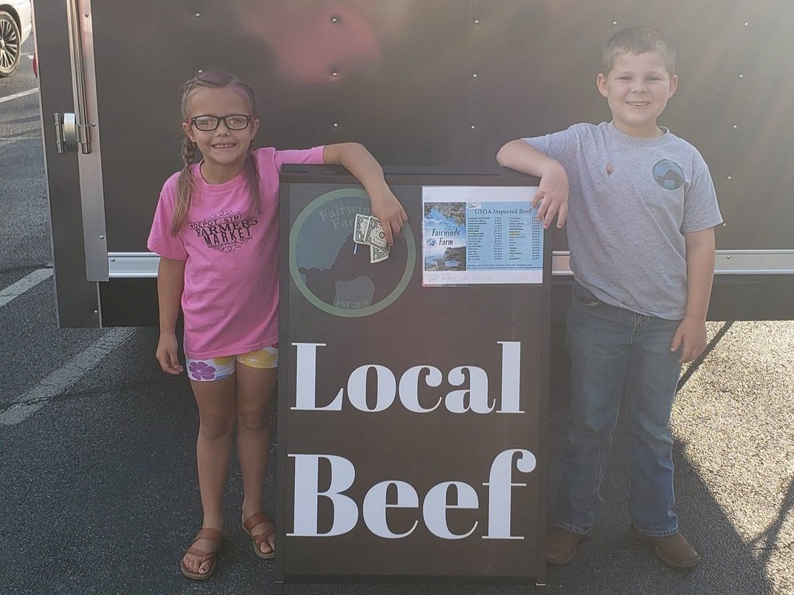 Two children stand in front of a vendor sign promoting local beef at a farmers' market. The girl, wearing a pink shirt and glasses, holds a dollar bill, while the boy in a gray shirt smiles beside her. The sign features the name "Fairwinds Farm" and a list of USDA inspected beef products.